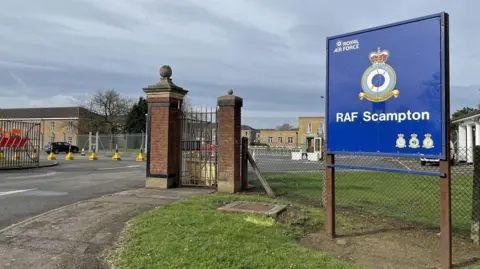 A blue RAF Scampton sign stands at the entrance to the site with an RAF logo and the badge of RAF Scampton. There are two brick gate posts at the entrance and buildings in the background. 