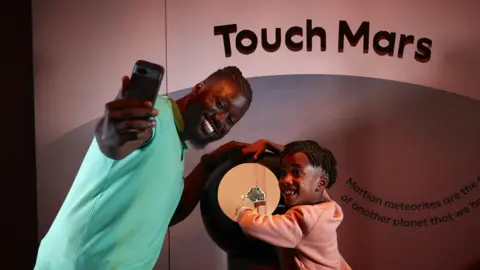 A man and a child pose for a selfie with an iPhone in front of a meteorite from Mars.
