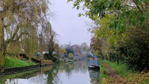 A narrowboat with smoke or stream rising from the top sits on a river surrounded by greenery and trees under grey clouds. 