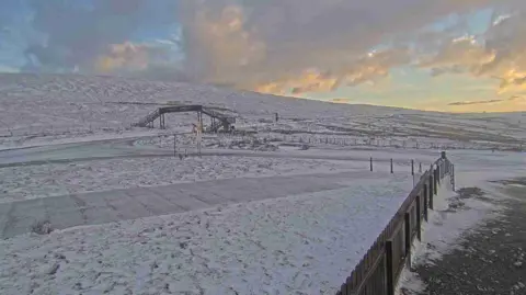 DoI A webcam view of the road at the Bungalow looking towards Onchan. There is a metal footbridge in the distance and the ground is covered with snow.