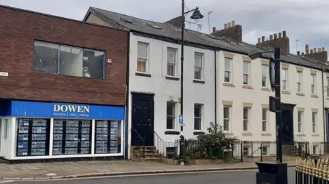 42 Frederick Street, Sunderland. A two-storey grey-white building with large, black front door and black detail around the windows, of which there are two on the ground floor and three on the first. Two skylights are visible on the roof. To its left is a modern, flat-roofed estate agent shop with a blue and yellow sign and a rectangular, plain window set in the brick-fronted upper floor.