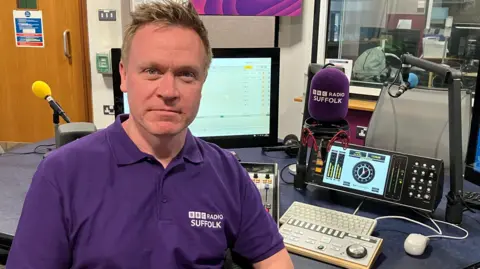 Graeme McLoughlin, who has short blonde hair, sits in a radio studio wearing a purple polo shirt with the "BBC Radio Suffolk" logo. He is surrounded by broadcasting equipment, including a purple microphone windshield with the same logo, a digital audio mixer, a computer monitor, and a keyboard.