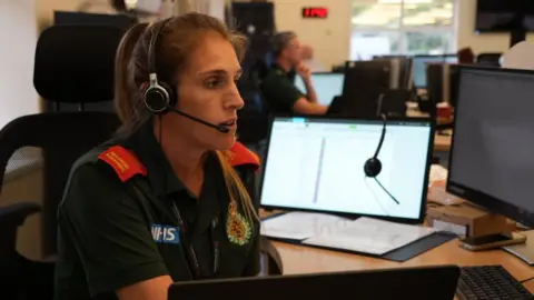 Shaun Whitmore/BBC Woman wearing green ambulance uniform with red epaulettes and the NHS logo sits at a desk and is looking at a screen. She has a headset and is talking to a microphone. She is in a control room with other similar personnel in the room. 