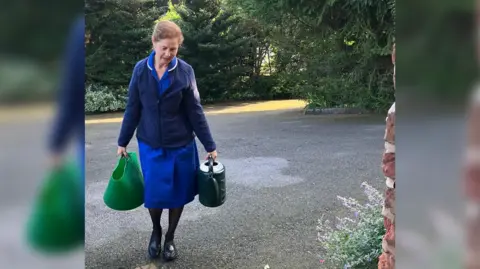 David Bradley A woman walking up a driveway, carrying a trug of water and a watering can. She's wearing a royal blue nurse's uniform with a dark blue coat over the top. 