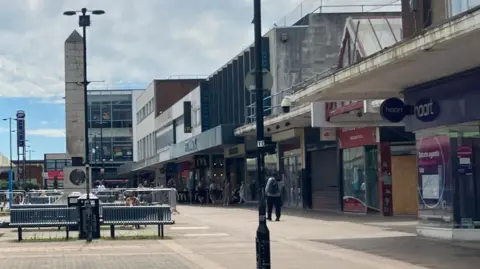 A shopping centre with benches in the middle and shops on the side, street lamp in the corner and people walking down the high street in broad daylight