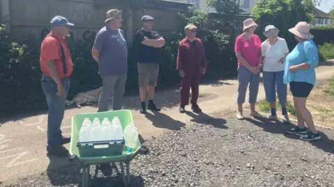 A group of seven people standing in a semi-circle talking to each other. They are all wearing hats because it is a sunny day. There is a green wheelbarrow full of plastic bottles with water in them.