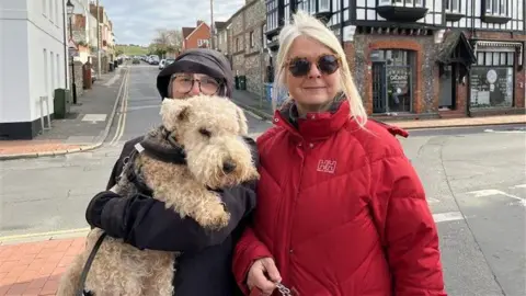 BBC/George Carden Dawn Jones (left) wearing a purple hat and coat covering her face with a brown dog and Siobhan Dodds (right) wearing a red coat, sunglasses and having blonde hair.