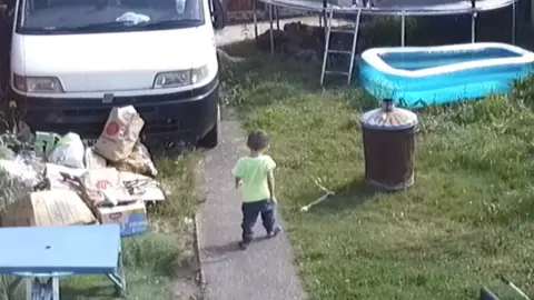 North Wales Police CCTV in the Ives' garden shows Ethan walking down a cement path, he is wearing socks, black trousers and a neon yellow t-shirt. To his left is grass and lots of rubbish, a blue plastic picnic table and a van. To his right is something, perhaps metal, lying in the grass next to a rusting bin. In the grass is a blow up paddling pool and a trampoline.
