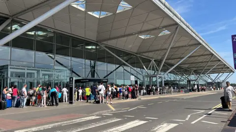 People queue outside Stansted airport departures area in the sunshine.