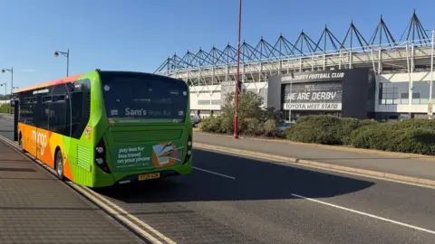Supplied A green and orange bus parked on the road outside a modern football stadium, with "WE ARE DERBY" printed above the entrance.