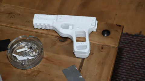 A small white 3D-printed handgun lying on a brown wood table next to a glass ashtray with stubbed out cigarettes in it
