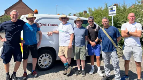 Seven men standing in front of a white van. They are all wearing different coloured t-shirts and a variety of shorts and trousers. There is a blue sky in the background.
