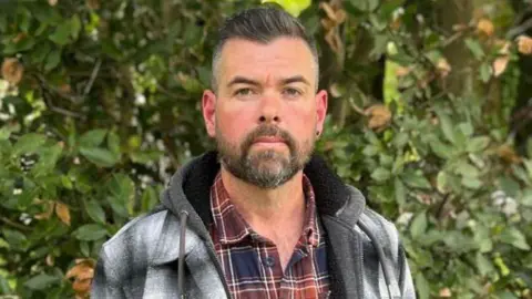 A man wearing a black and white checked jacket and red and blue checked shirt stands in front of shrubbery. He has a short beard, dark in colourand short dark hair. He is looking directly at the photographer.