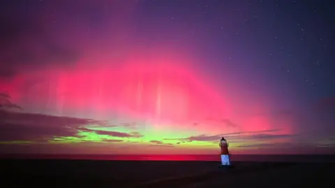 Landscape shot of the northern lights from the Point of Ayre. A lighthouse can be seen looking over the coast whilst the sky is lit in bright pinks, greens and purple.