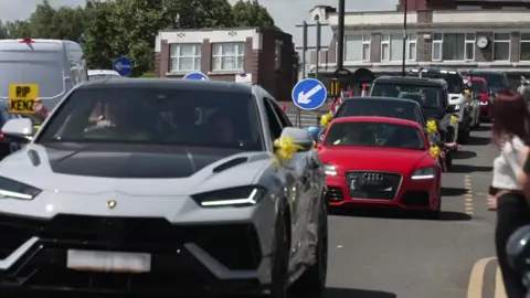 Cars with yellow ribbons on their wing mirrors travel down a road.