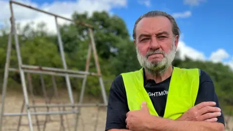 Christopher "Chip" Thorogood, standing in front of several metal A-frames. Chip wears a black T-shirt with white lettering, and a yellow high-vis on top. Chip has dark hair brushed back and a dark grey beard.