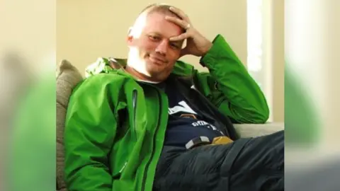 London Ambulance Service Mark Pell sits on a fold-out sofa. He is wearing a light green waterproof jacket open in the front and is smiling, placing one hand on the top of his head and forehead 