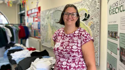 Bex smiling at the camera in front of a table filled with folded school uniform items.