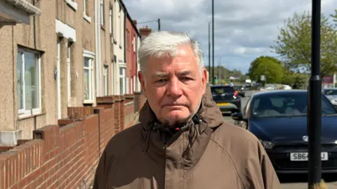 BBC/Jim Scott Bill McGlen is wearing a brown zipped up jacket while standing in front of homes and parked cars on Chester Road. In the background are other vehicles and trees which are opposite Herrington Country Park.