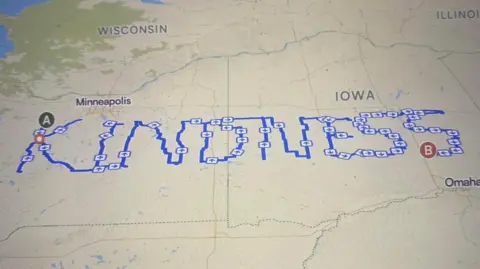 Maplibre/komoot/Map data: OpenStreetMap Contributors A digital map on the United States with a route marked out in a blue line. The route spells out KINDNESS and starts in the Minneapolis area and ends near Omaha.