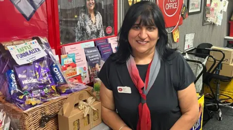 Roopie Gill standing behind a Post Office counter beside a raffle fundraiser display featuring a wicker hamper, chocolates, biscuits, and gift items. A large poster in the background and displays a photo of a staff member. The shop interior includes Post Office signage, product brochures, and community noticeboards.