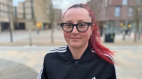 Dani Cattani looking into camera with buildings behind her in the background. She has long red hair, tied up, black rounded glasses and is wearing a black Adidas jumper with white trim.