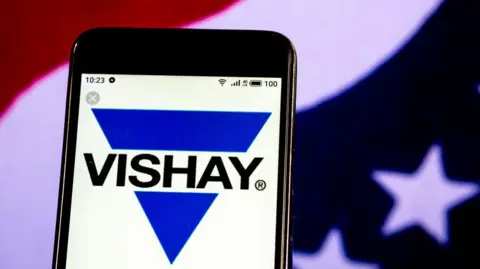 The Vishay Intertechnology Semiconductor manufacturing company logo displayed on a smartphone. The logo is a blue triangle pointing downwards on top of a white background. The triangle is broken across the middle with the word VISHAY in black capital letters. A blurred American flag is in the background behind the phone.