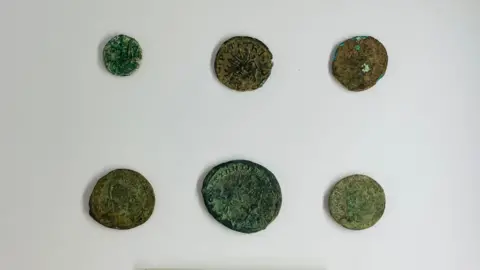 AC Archaeology and Heritage Six Roman coins on a white background all of various different shapes and sizes.