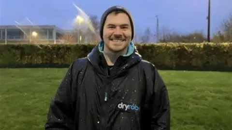 Group member James smiling for the camera. He is wearing a dark coat and a hat. He is wet from the rain. A green area could be seen behind him.