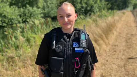 Sophie Hyde, standing in a field, smiling and looking straight at the camera. She has long blonde hair tied back, and is wearing a black police uniform. 