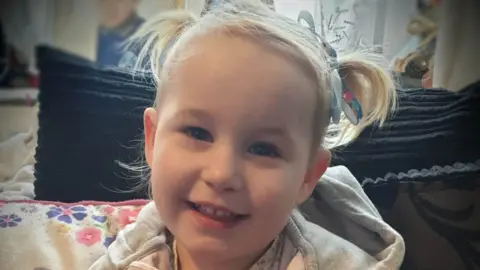 PA Media Lola James smiles for the camera, she has staright blone hair worn in pigtails and is wearing a grey hoodie