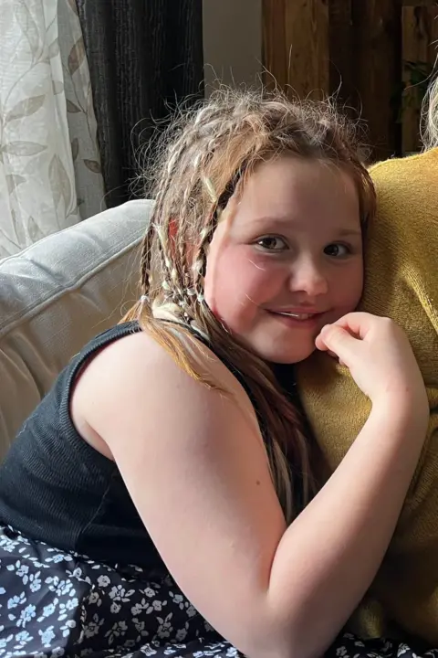 Family handout. Mya, A young girl aged about 8 is smiling to the camera. She is leaning on her mum's shoulder who is out of the frame. She has braids in her brown hair and she's wearing a summer dress. 
