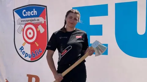 Karolina Urbanova Karolina Urbanova squints and smiles into the sun while holding an axe and wearing her hair in two braids