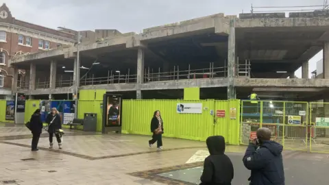 The site of the new Community Diagnostic Centre with green fencing. Part of the structure of the old Broadmarsh Centre is being retained for the new building and is visible behind the fence with people walking in front.