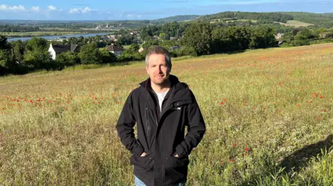 Reserve Manager Ben Sweeney stands in a field full of wild flowers on a hillside looking down onto the River Medway