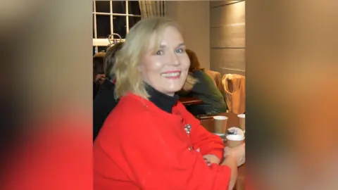 A woman wearing a red top smiles at the camera. She has short blonde hair and is sitting a table holding a paper cup.