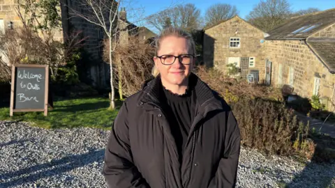 Nathan Turvey/BBC Lucy Fleming, who works at a wedding venue and cafe at Otley Chevin, wearing glasses and a black coat with stone built buildings and a bush in the background. 