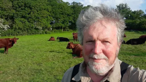 A man with windswept grey hair and a grey beard, wearing a brown shirt, stands in a field with several brown cows and one black cow behind him.