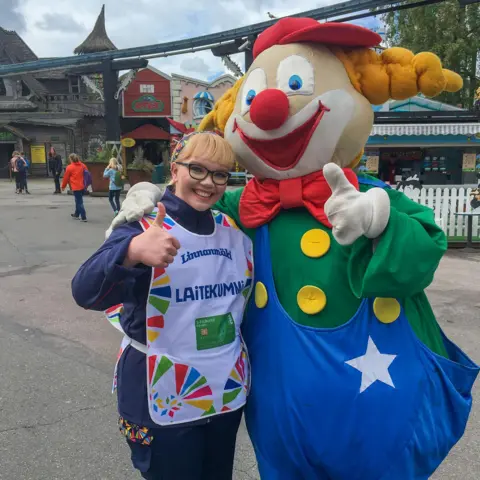 Meri-Tuuli Auer Meri-Tuuli Auer poses with a clown mascot at an amusement park. They are giving a thumbs up to the camera.