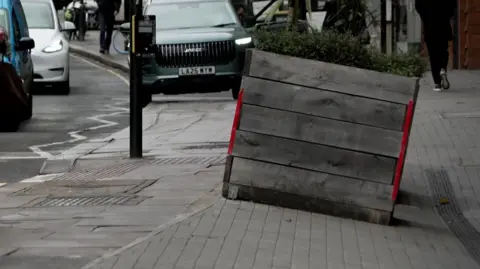 The sloping pavement with a big planter on it. The background includes cars and pedestrians.