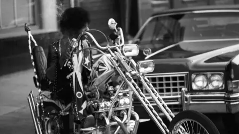 Getty Images Sly Stone rides a large elaborate chopper style motorbike in a black and white image. He has a leather jacket on with a large white star on the collar.