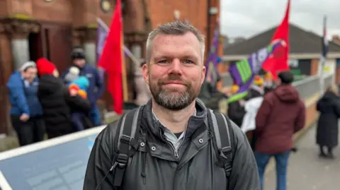 PA Media Gerry Carroll is standing outside a red brick building with a group of people who are taking part in a protest. He is wearing a dark green coat and a rucksack with black handles. 