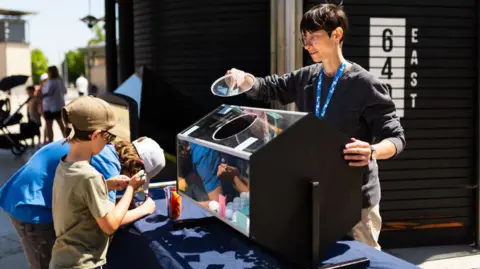 We The Curious A member of staff is standing beside a tombola machine, holding a round glass lid away from the opening, He is looking at two boys who are writing on pieces of paper to add to the tombola. The exhibit is in an outside space in sunshine