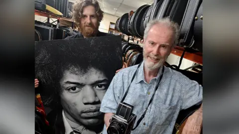 Gardiner Houlgate Two men, one with a camera and one with a large picture of Jimi Hendrix, stood inside a building.