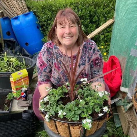 Christine Rutter Christine Rutter. She has shoulder-length brown hair and is holding out a basket containing green leafy plants and a taller red-leafed plant.