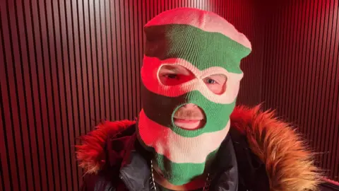 Rapper Durt Burd. He is wearing a green and white stripped balaclava, black coat with fur lined hood and gold chain. He is standing in a recording studio with red lighting.