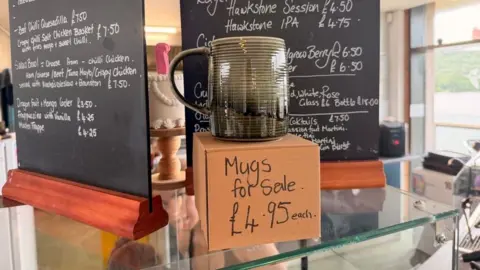 A green mug on a brown box with 'mugs for sale £4.95 each' on the box.