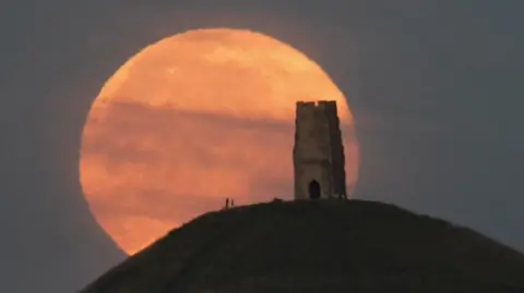 A reddish looking full moon behind a structure on a hill