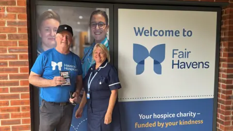 Supplied Alan stands wearing a blue T-shirt, with the words HAVENS HOSPICES, while next to a nurse who is also standing. They are in front of some branding, with the words FAIR HAVENS printed on a window.