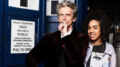 BBC Studios Peter Capaldi, with wavy grey hair wearing a burgundy velvet blazer and white shirt and holding his hand over his face in a puzzled manner with a frowning expression on his face, is on the left of the picture. Beside him is a woman in a black and white t-shirt and denim overshirt. She has black afro hair and is smiling at the camera. Behind them is a blue phone box, known as the Tardis.